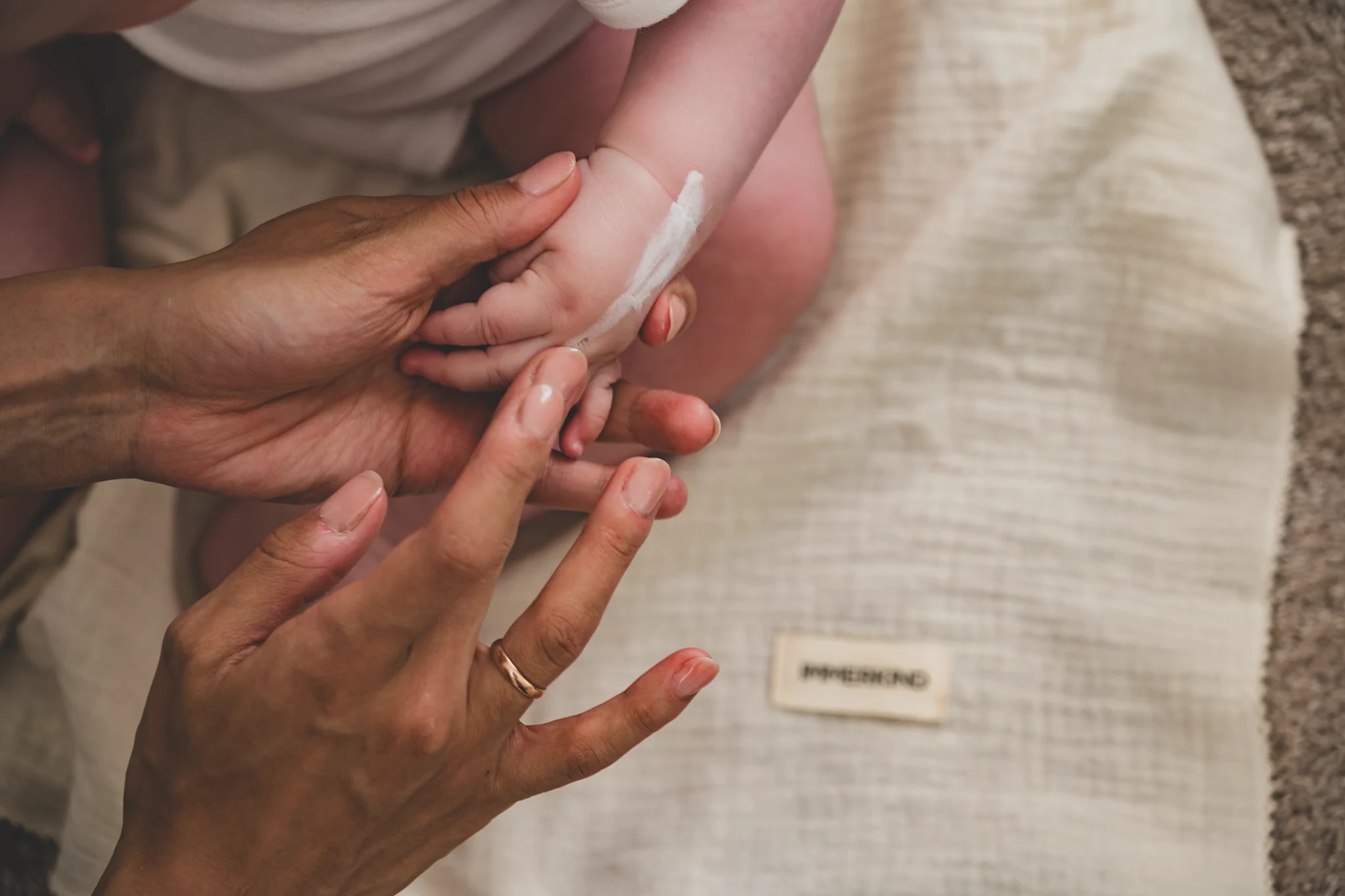 Erwachsene Hand trägt Lotion auf den Arm eines Babys auf, während es auf einem beigen Stoff liegt.